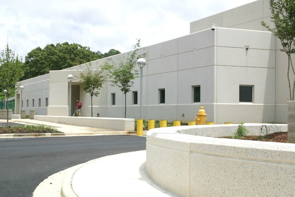 Exterior of a light-colored building with small windows, a curved concrete barrier, and trees. A person stands by the entrance.