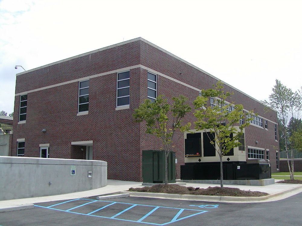 Brick building with rectangular windows; trees and paved parking area in front.