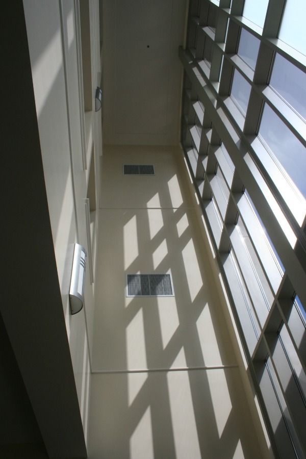 Interior shot of a tall, light-filled atrium with windows. Sunlight casts shadows on the beige walls and the metal framework.