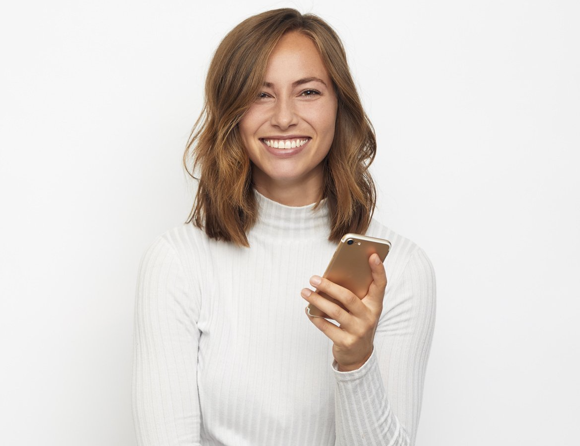 Eine Frau in einem weißen Pullover lächelt und hält ein Mobiltelefon in der Hand.