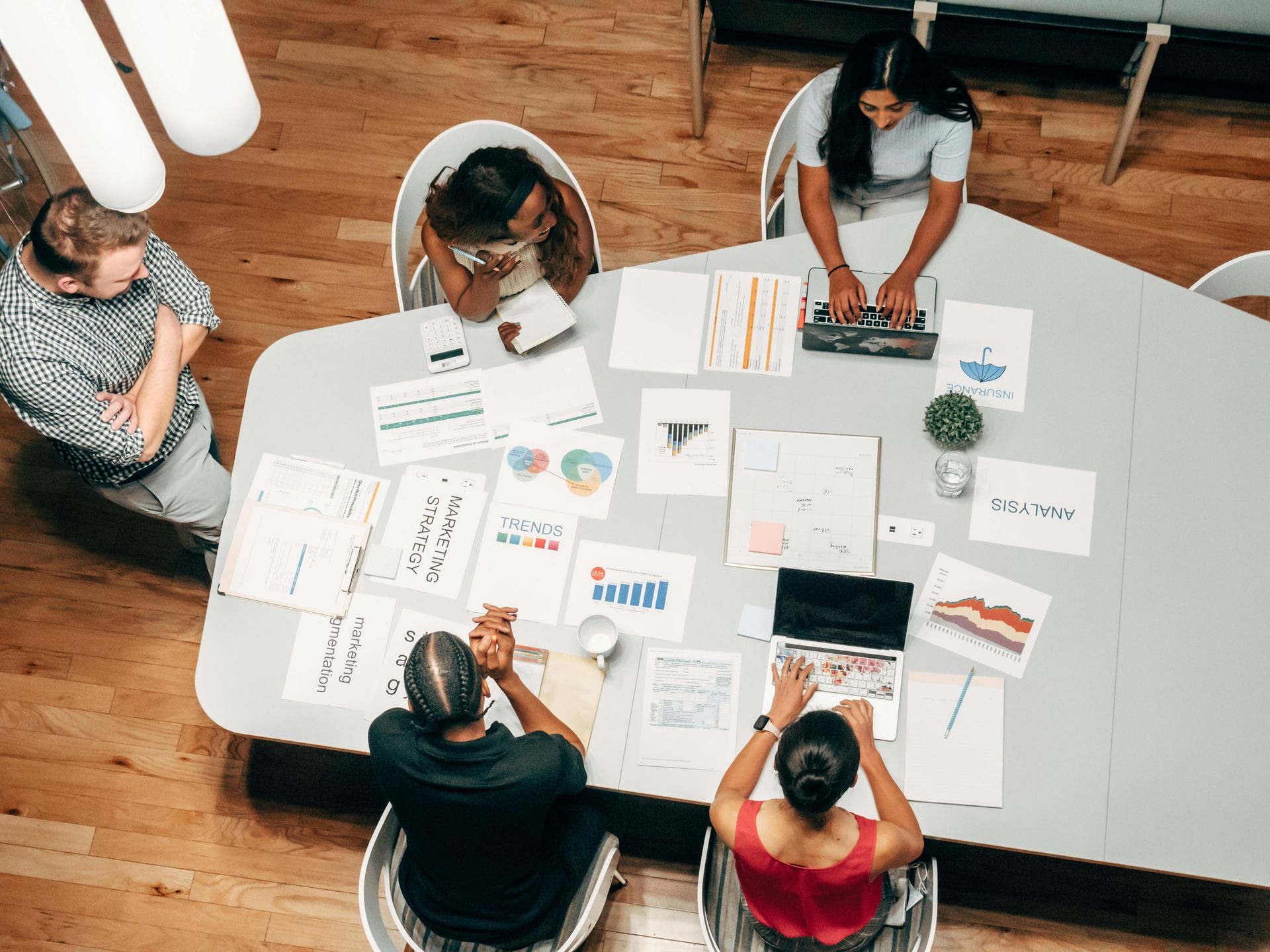 Group of diverse people collaborating around a table, discussing documents and using laptops in a modern office.