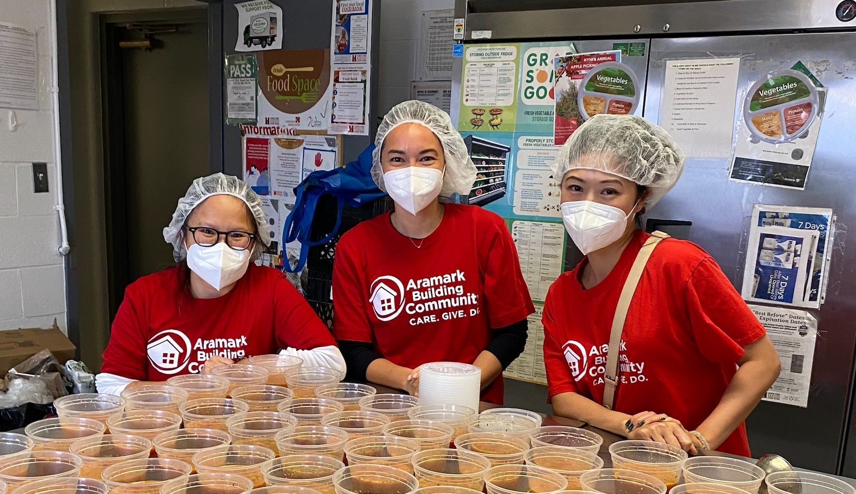 Three women wearing face masks and hair nets are standing in front of a table filled with bowls of food.