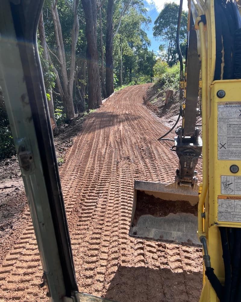 An Excavator Scoops Dirt on a Red Dirt Road Through a Forest — Heller Excavation & Tree Services in Mullumbimby, NSW