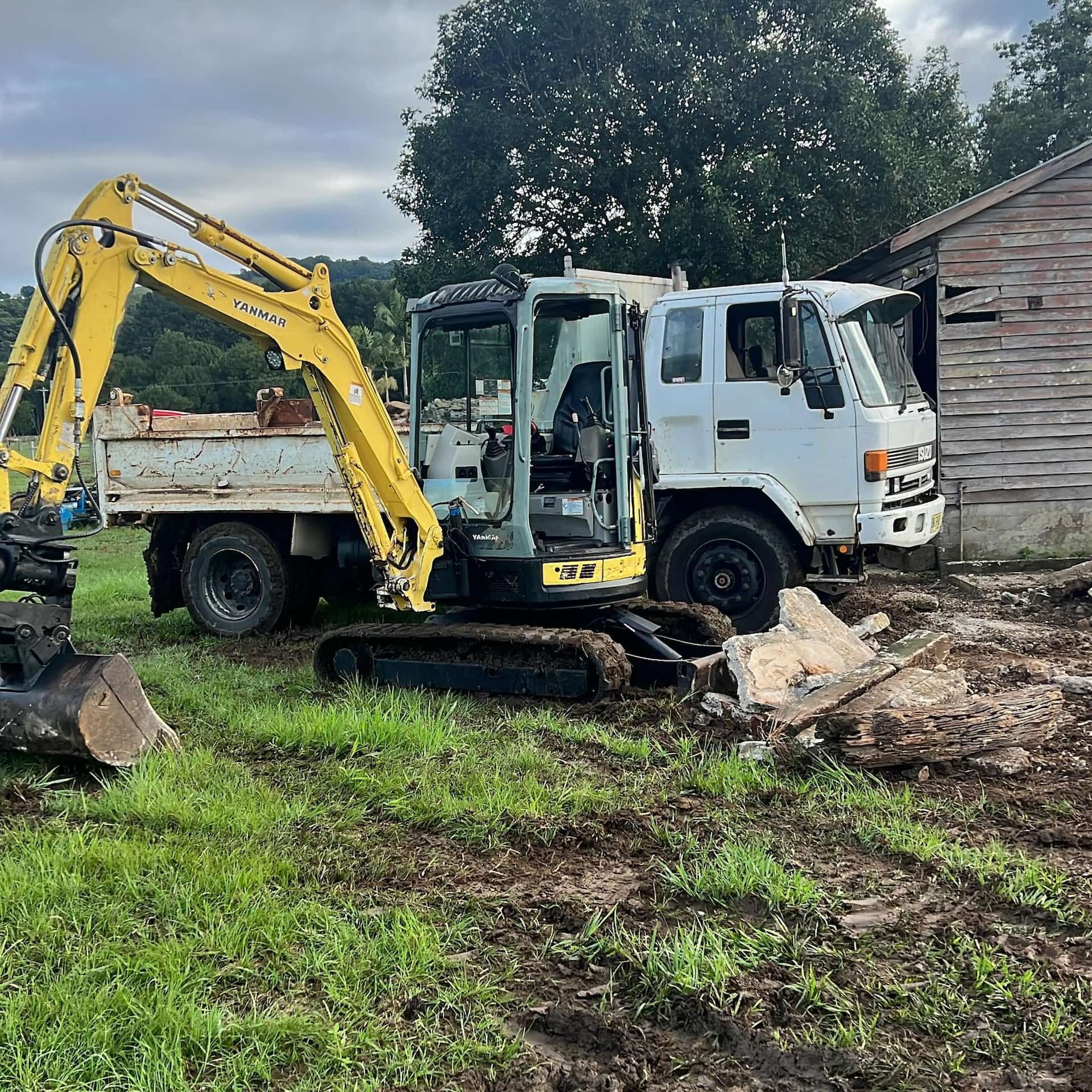 A yellow excavator and white truck next to a wooden shed on a grassy field.