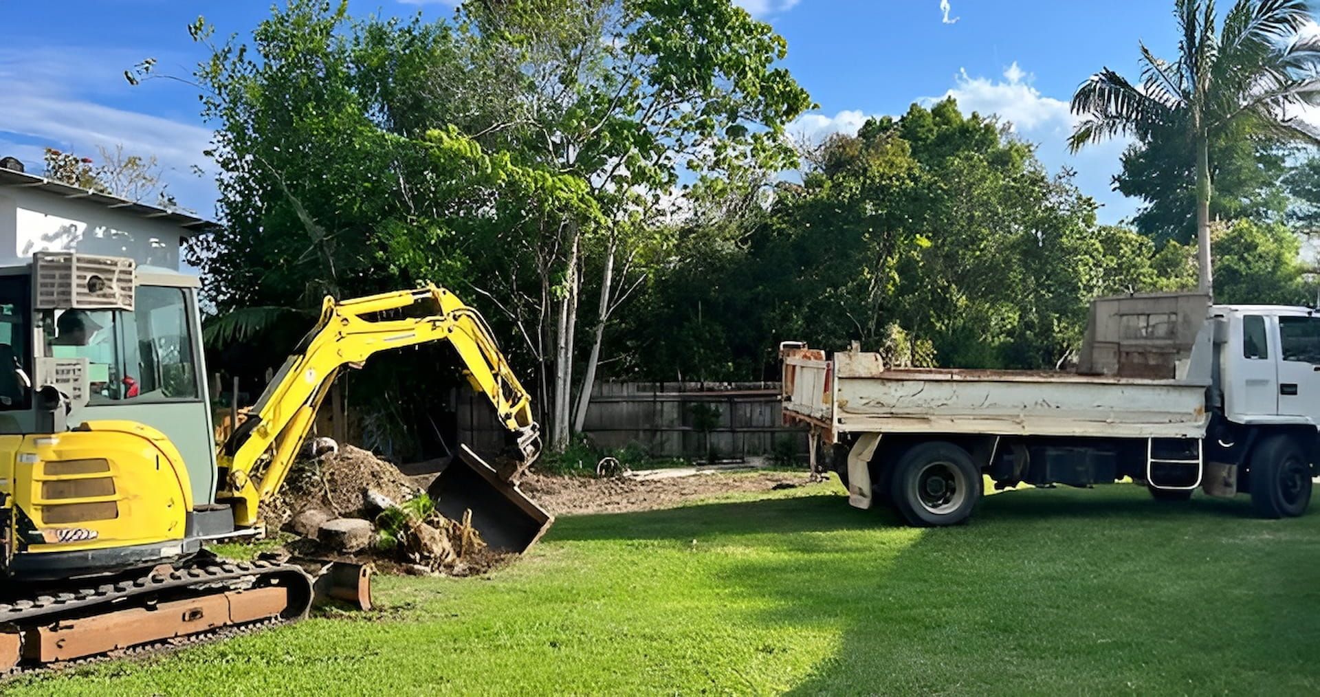 Mini excavator loading a dump truck with dirt on a grassy lawn. Yellow and white machinery, sunny day.