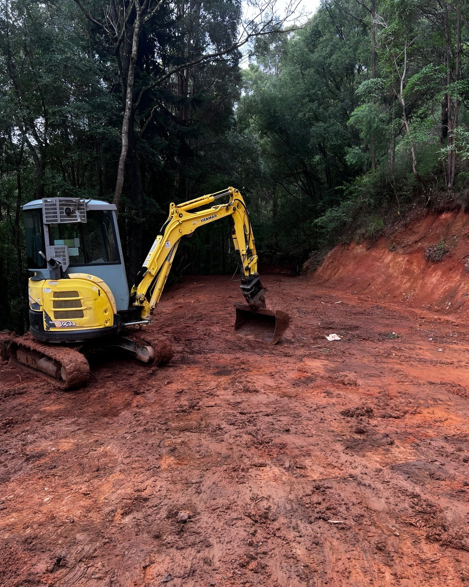 Yellow excavator on a muddy red dirt road in a forest.