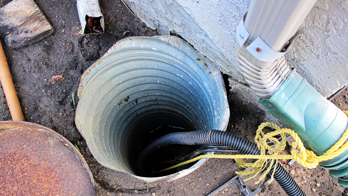 Open utility hole with a black hose, yellow rope, and PVC pipe beside a concrete wall.