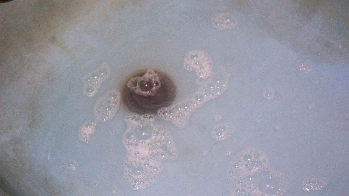 Small dark drain plug in pale blue water with soap bubbles around it