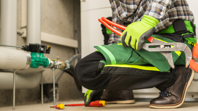 Worker kneeling beside industrial pipes, wearing green safety vest and gloves while handling tools.