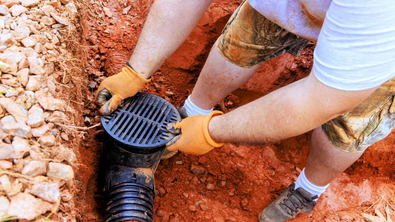 Hands install a black drain grate in red soil next to a corrugated pipe.