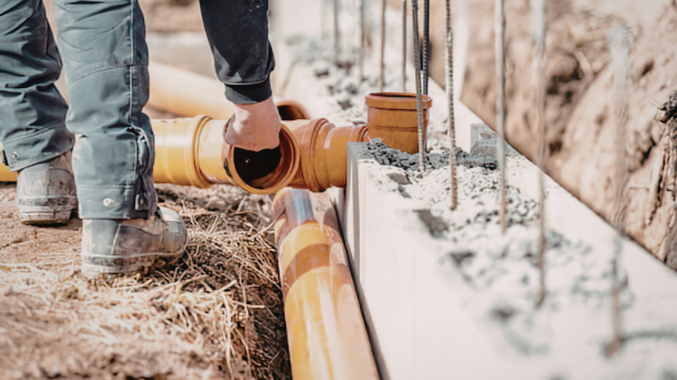 Worker laying orange drainage pipe beside a concrete foundation at a construction site
