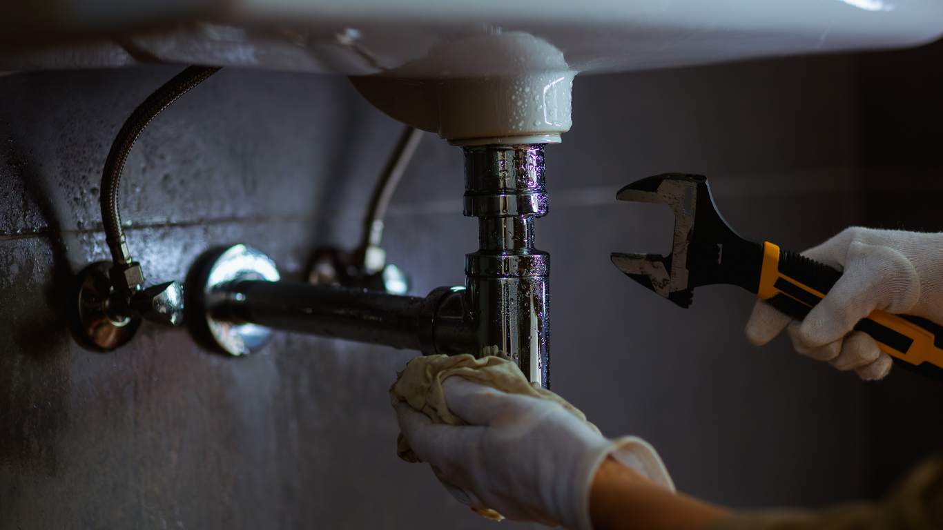 Hands repairing a sink drain under a bathroom sink with a wrench.