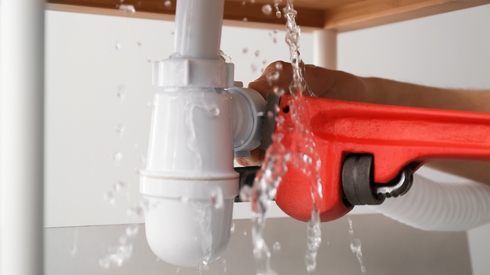 Leaking under-sink pipe with water spraying onto a red bucket and hose