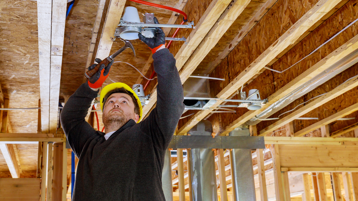 Construction worker using a red power tool while standing beneath exposed wooden ceiling beams.