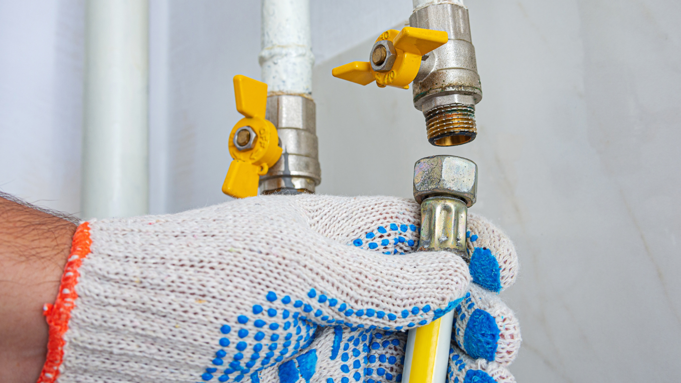 Gloved hand connecting a hose to a brass faucet with yellow shutoff valves on a wall