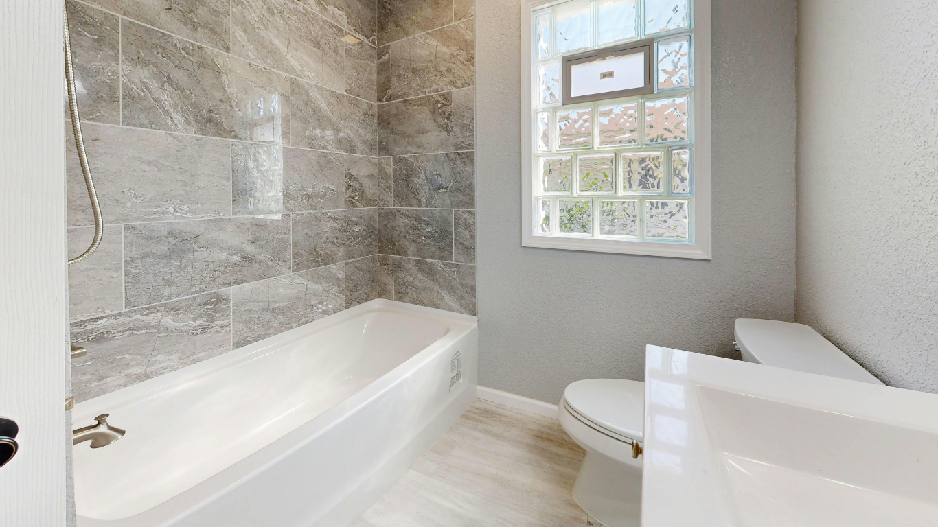 Bright bathroom with white tub, gray tile walls, and a window above the toilet