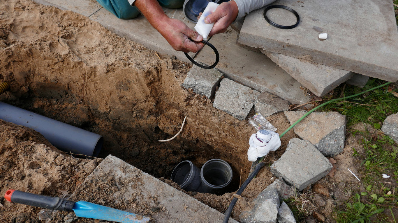 Hands working on an outdoor trench, fitting black cables and gray pipes near concrete pavers.
