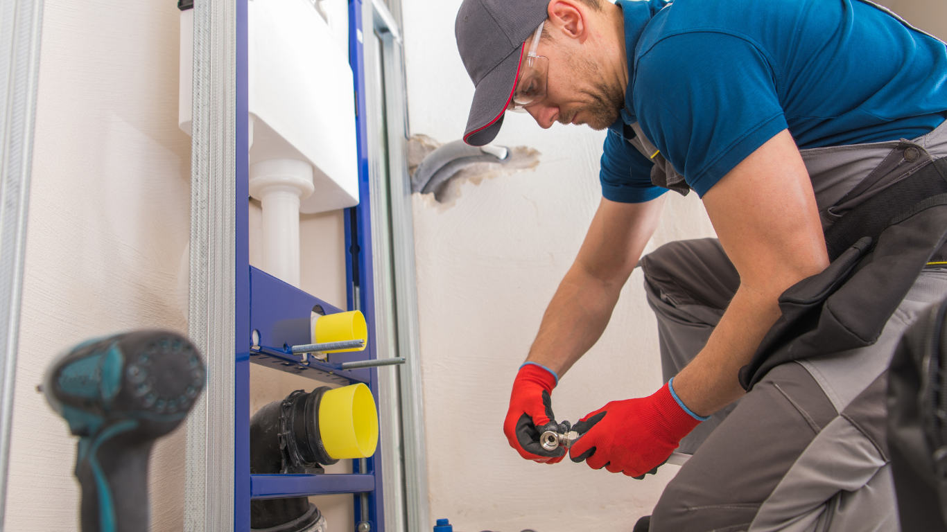 Technician in blue shirt and red gloves installing plumbing under a sink, using tools in a bathroom.