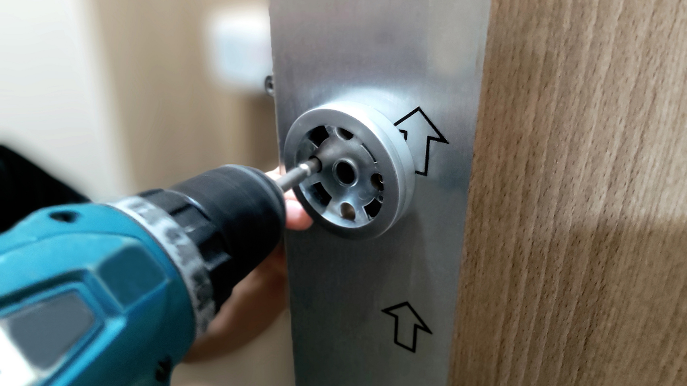 Blue cordless drill tightening a silver furniture connector on a wooden panel