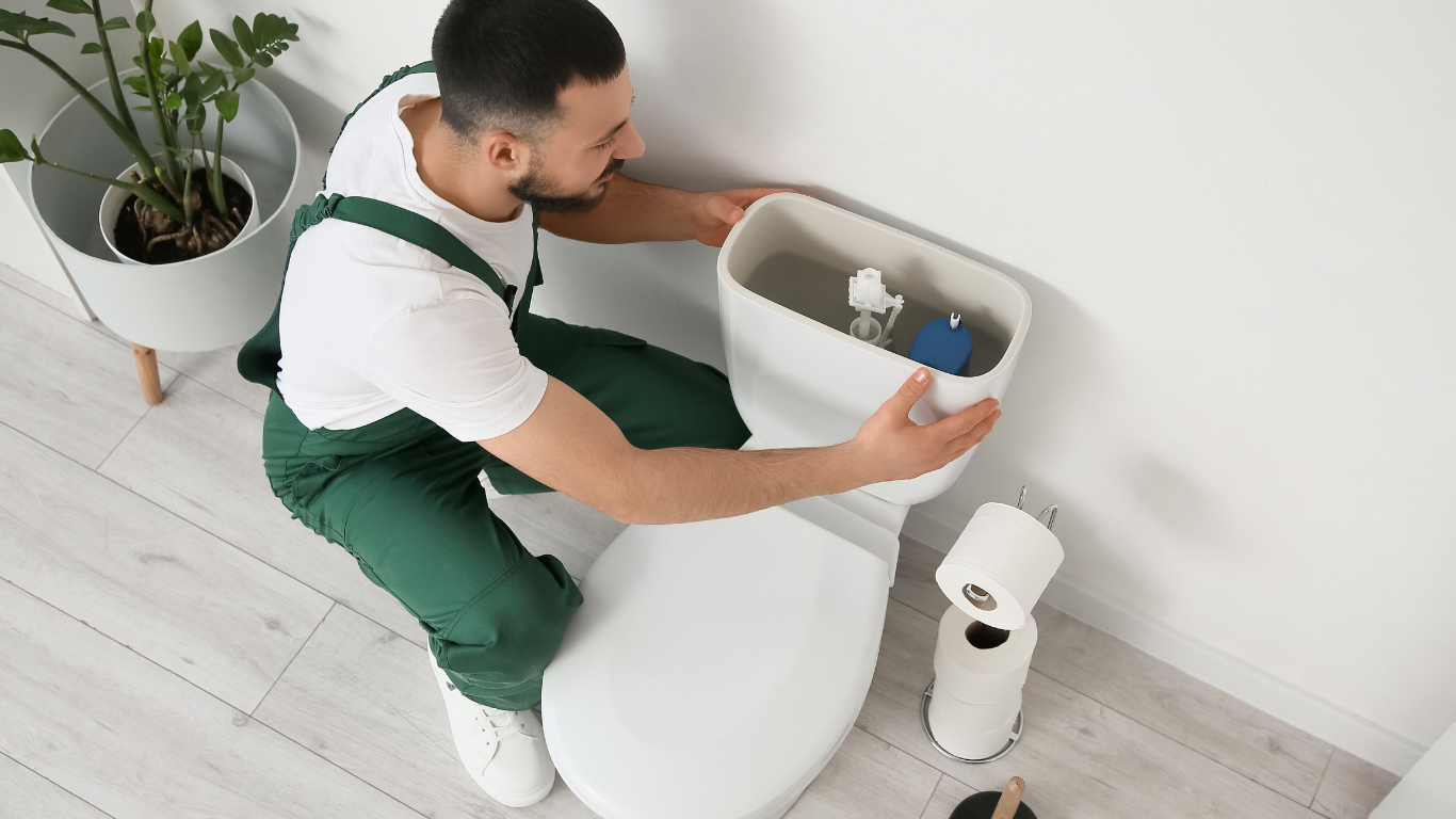 Plumber in green overalls installing a toilet tank in a bright bathroom