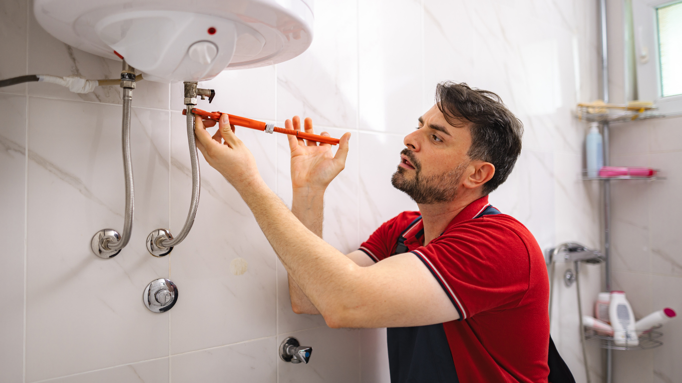 Technician repairing a white water heater with a red power tool in a tiled bathroom