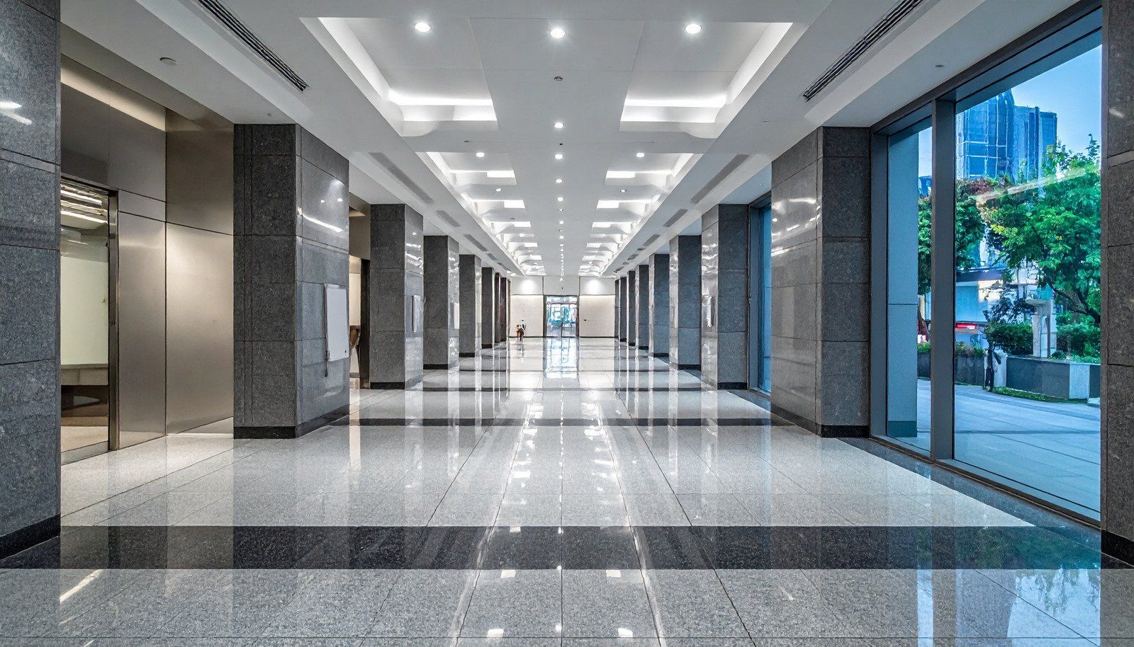 Long hallway in a modern building with marble floors and columns, natural light from large windows.