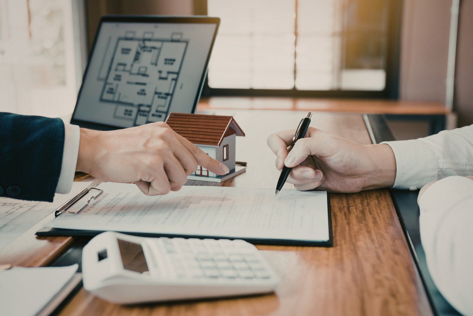 Two people signing a real estate contract with a house model, blueprint, and calculator on the desk.