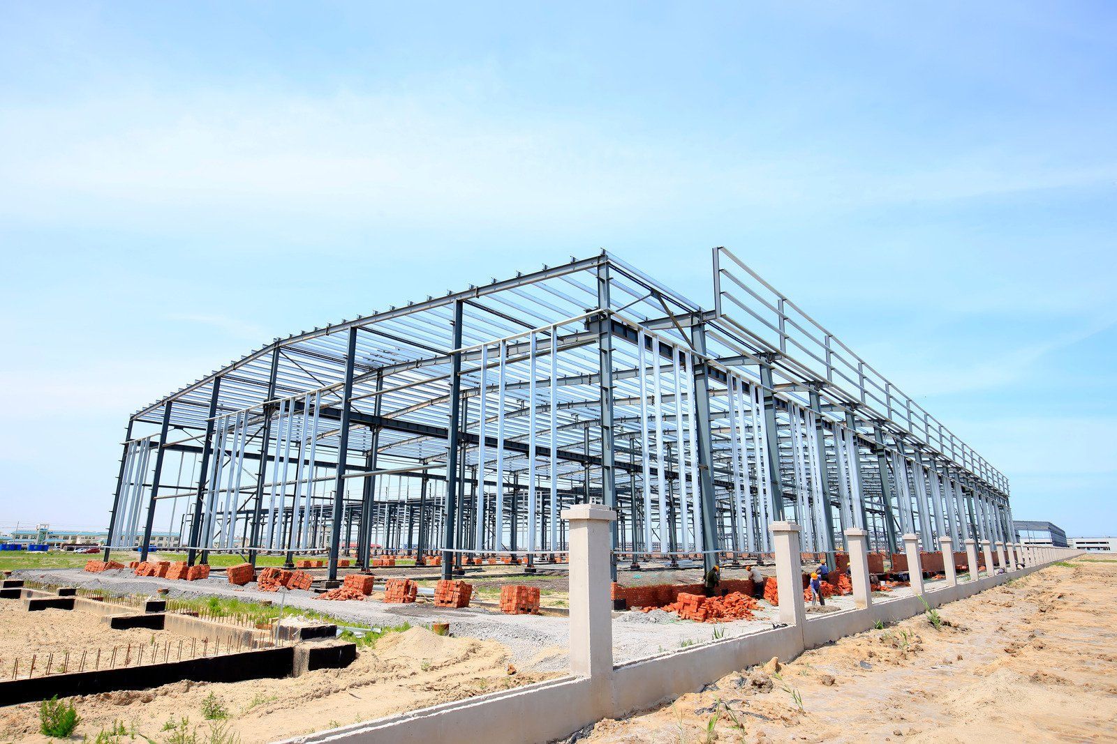 Steel frame of a large industrial building under construction, set on a concrete foundation, against a clear blue sky.