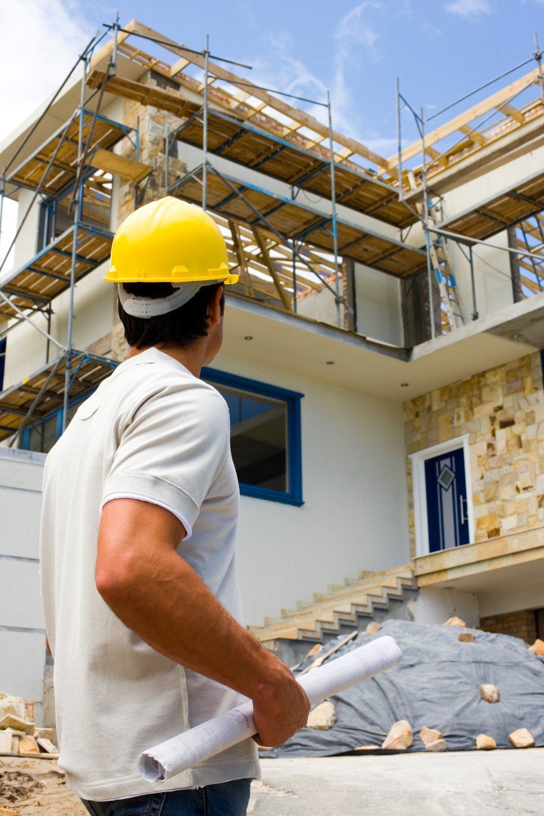 Construction worker in yellow hard hat looks at a house under construction, holding blueprints.