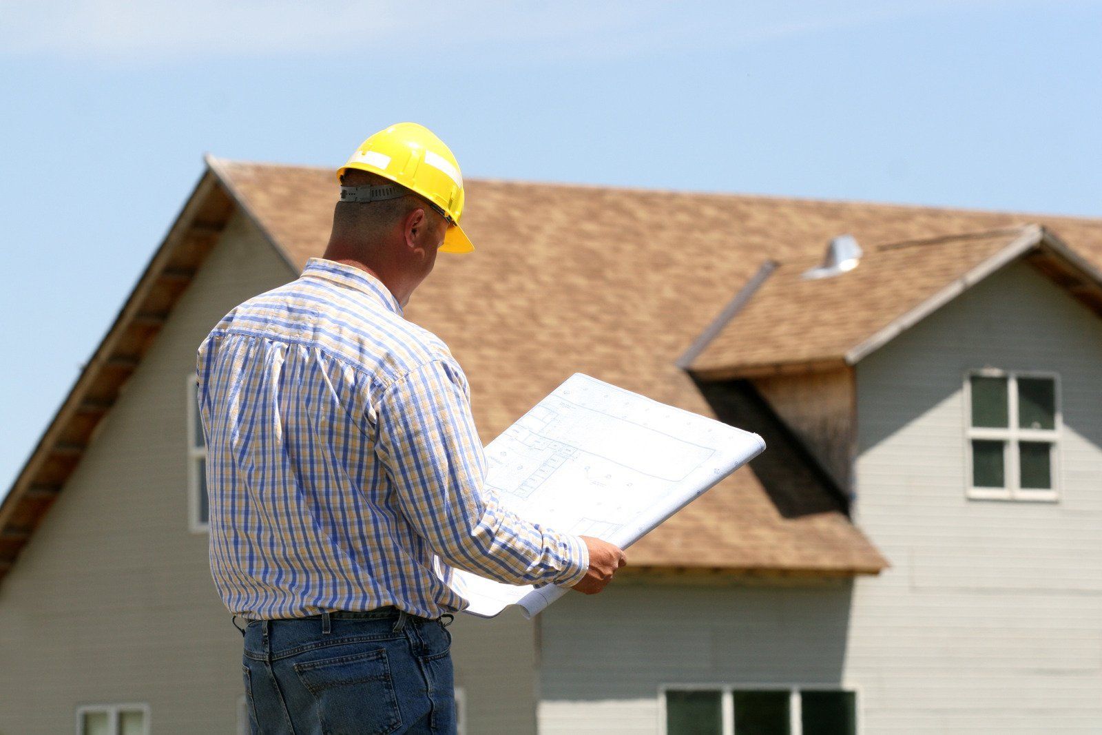 Construction worker wearing a yellow hard hat examines blueprints in front of a house.