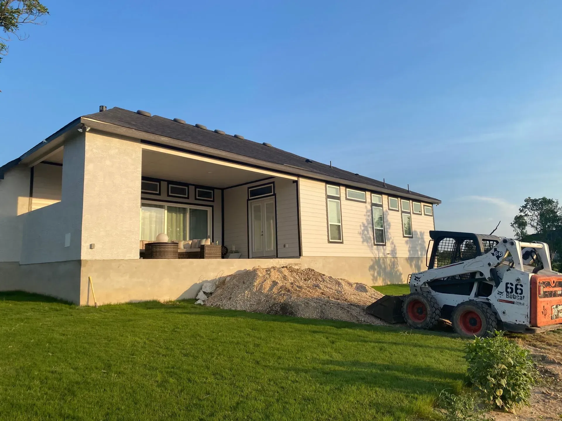 A white house with a covered patio, a pile of gravel, and a Bobcat on a grassy lawn under a blue sky.