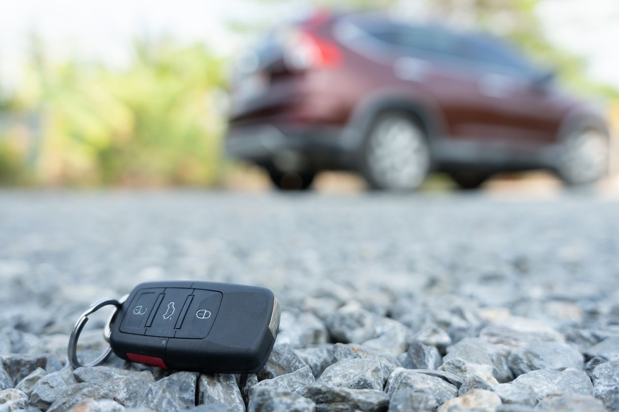 Car key fob on gravel, with a blurred brown SUV in the background.