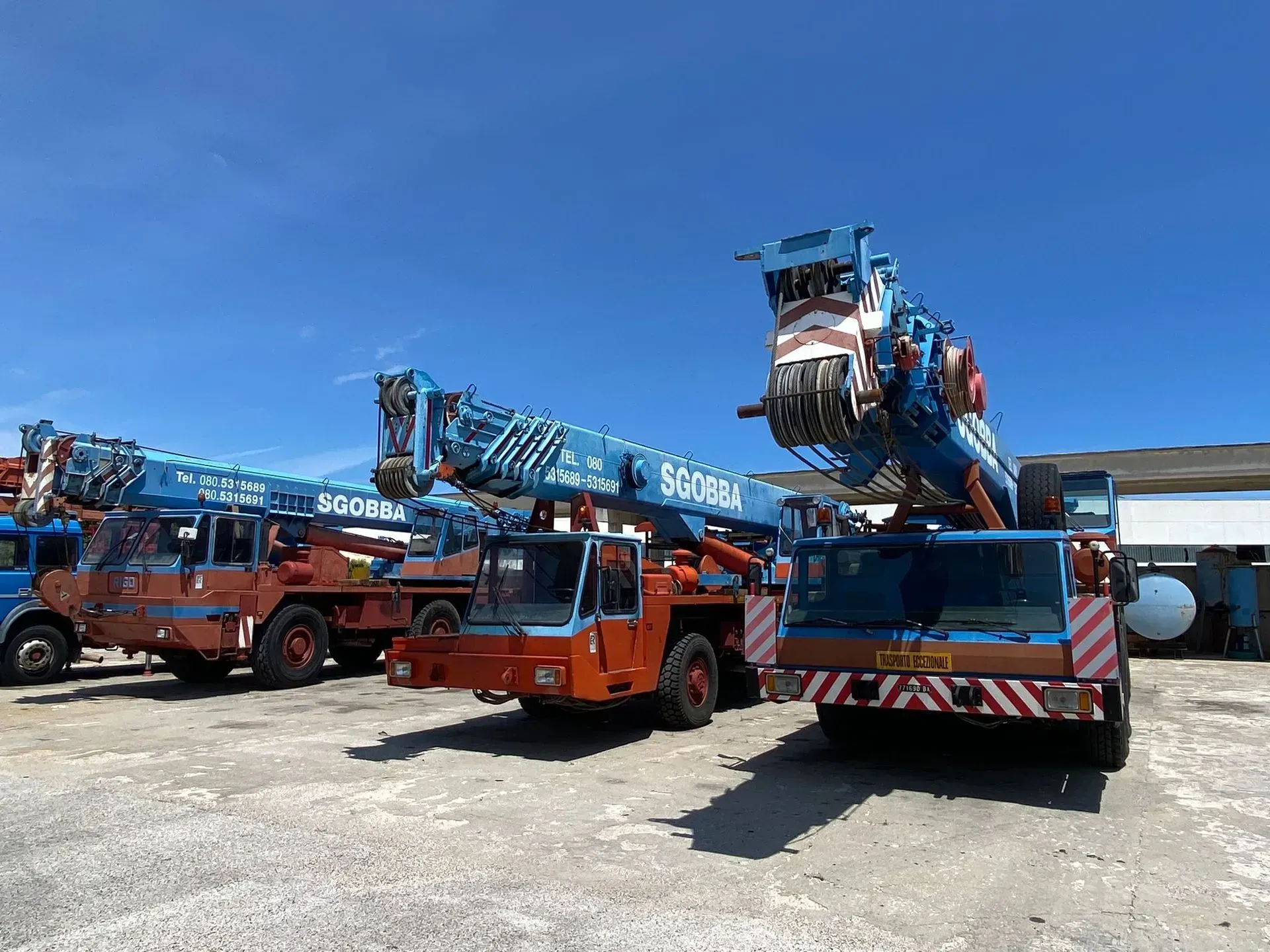 Tre gru arancioni e blu parcheggiate in un cantiere soleggiato sotto un cielo azzurro e limpido
