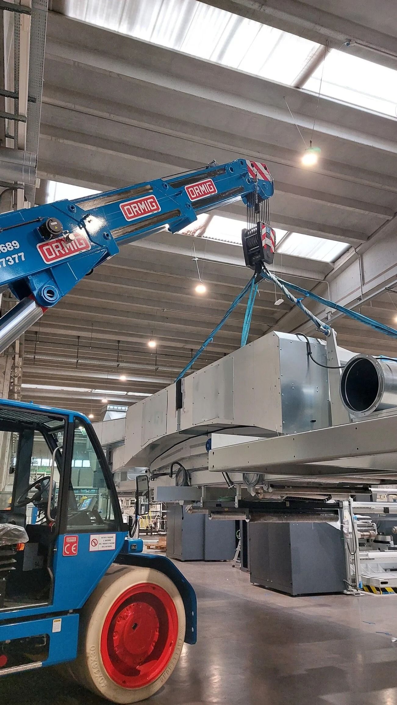 Un carrello elevatore blu solleva una grande trave metallica all'interno di un magazzino industriale.