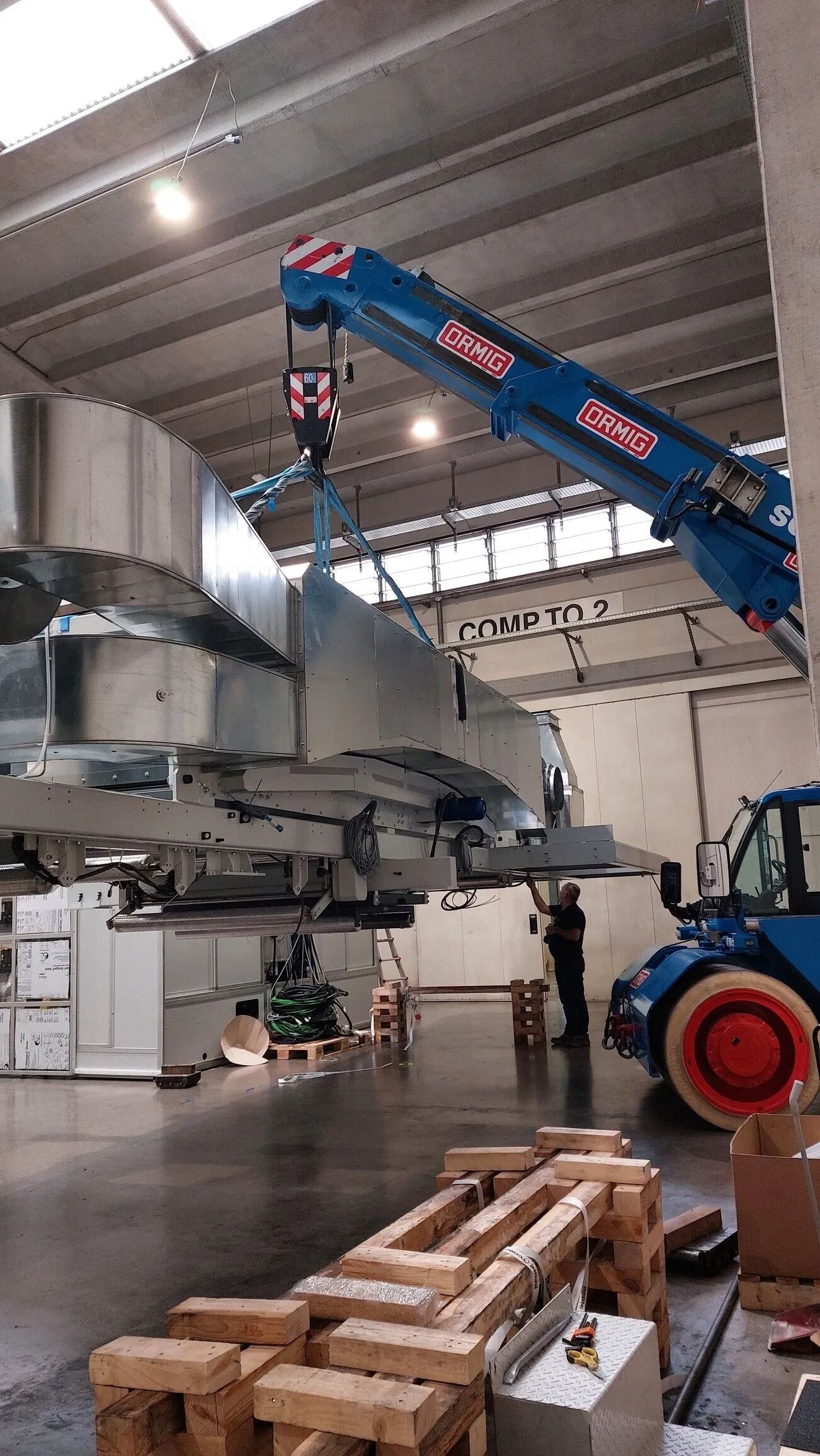 Una gru blu solleva un grande componente metallico di un aereo all'interno di un'officina industriale.