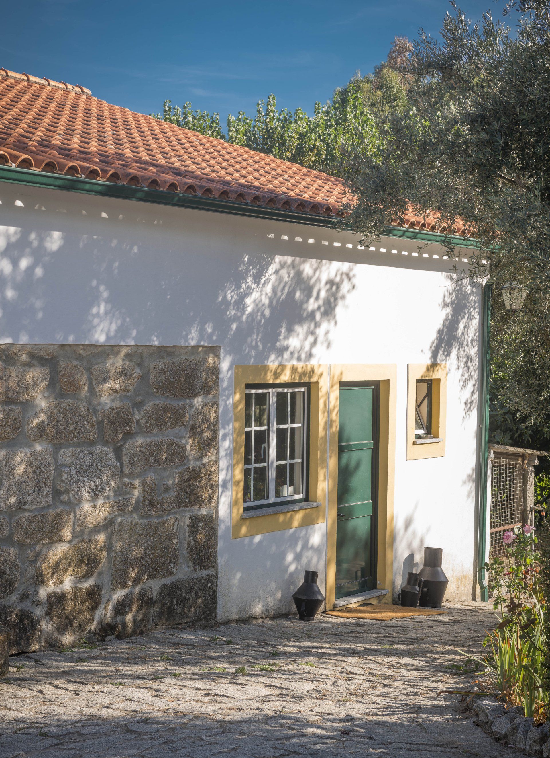 White building with a red-tiled roof, stone wall, and green door. Yellow trim. Outdoors, sunny.