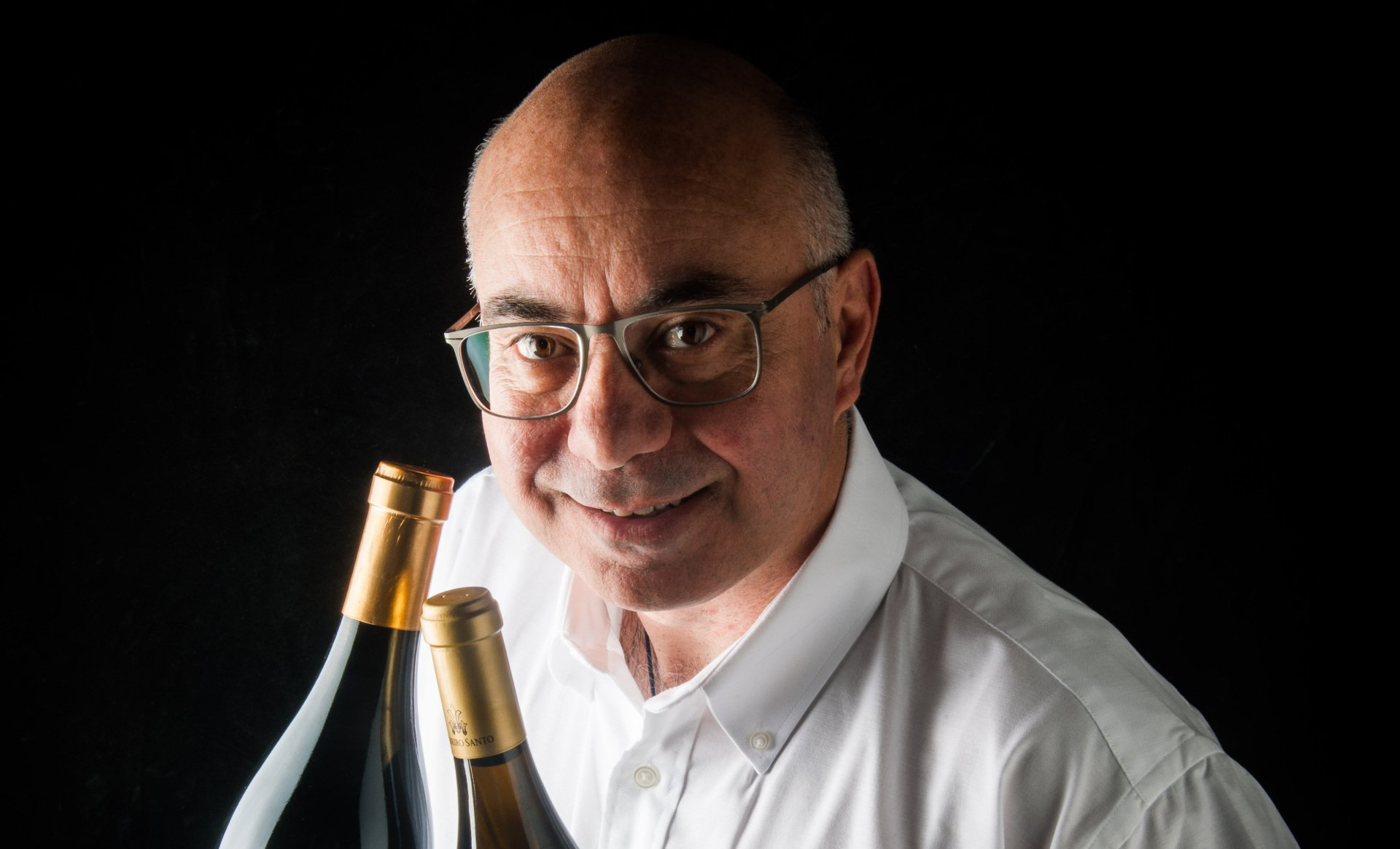 Man holding two wine bottles, smiling, wearing glasses and a white shirt against a black background.