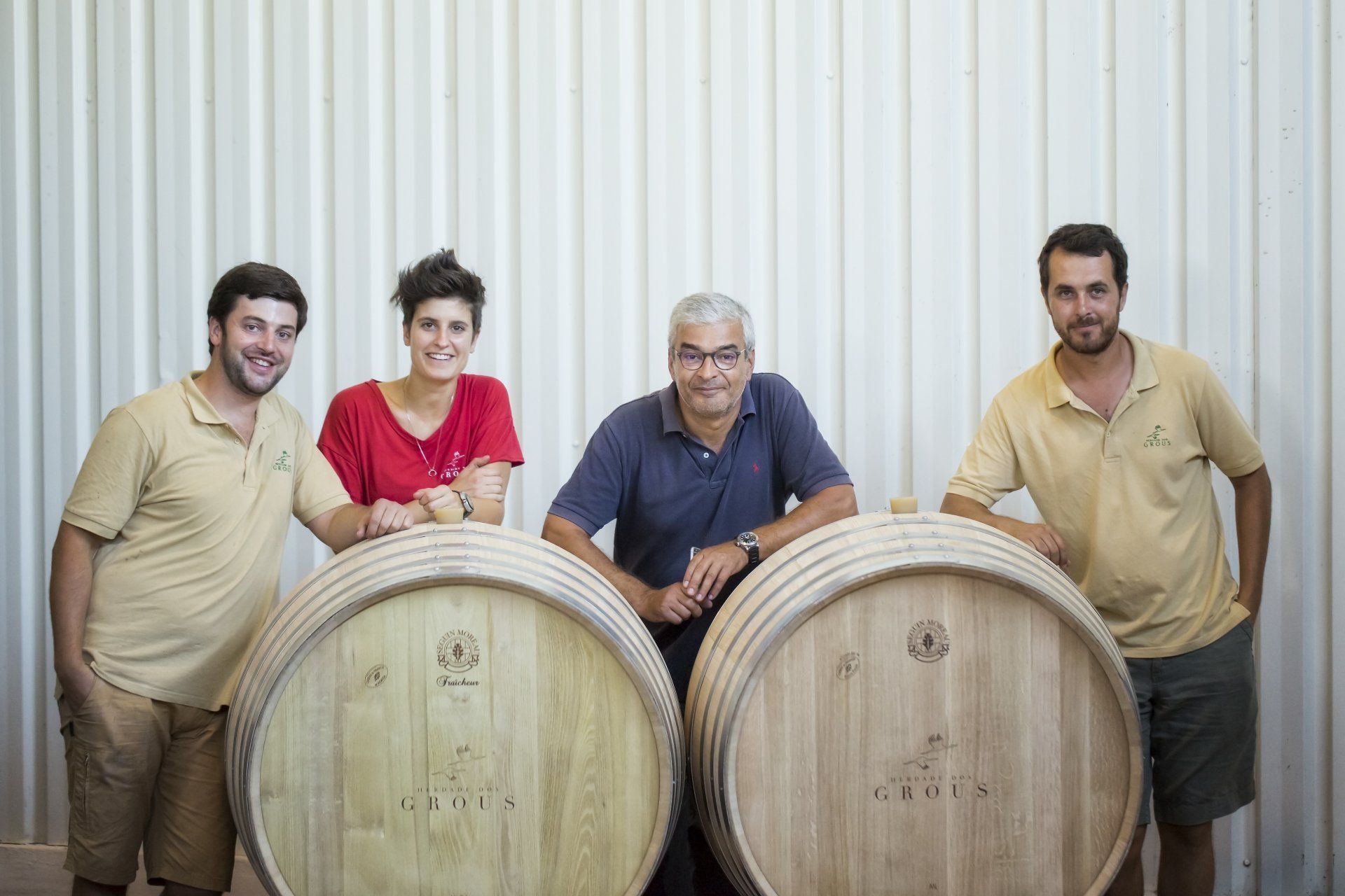 Four people in a winery pose with wooden barrels. They lean on the barrels, smiling. Pale walls are the backdrop.