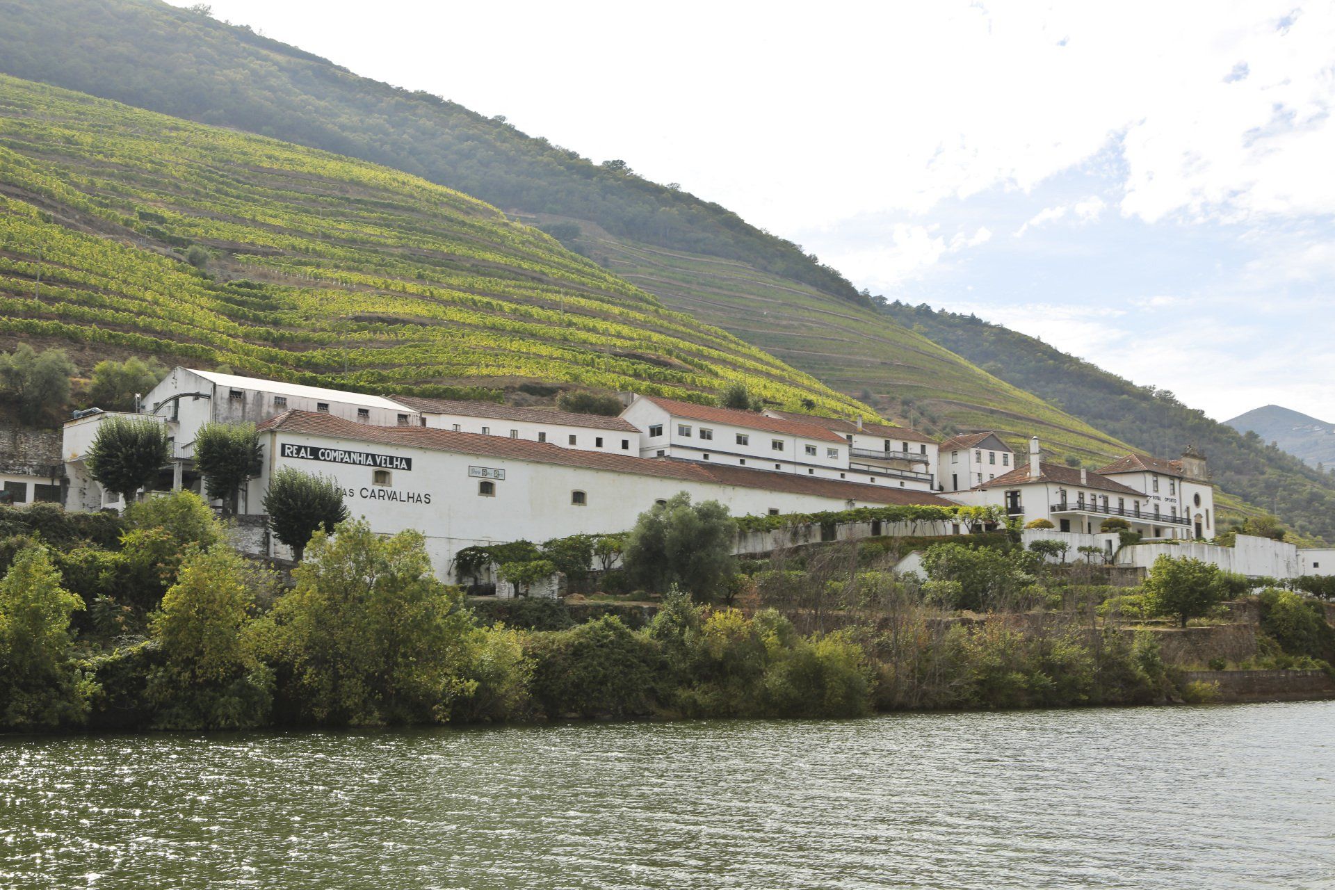 White building with red roof tiles on a riverbank, surrounded by green vineyards on a hillside.
