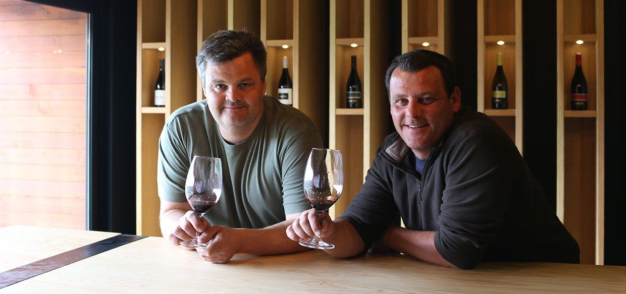 Two men holding wine glasses, smiling at the camera, seated at a wooden bar. Wine bottles in cubby shelves.