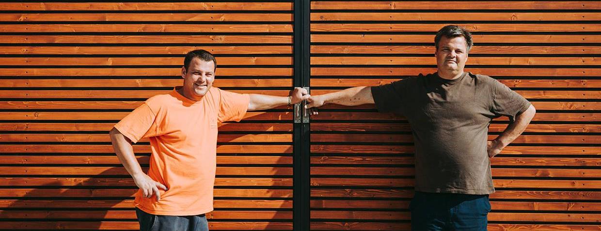 Two men standing on either side of wooden doors, hands on the handles. One wears orange, the other brown.