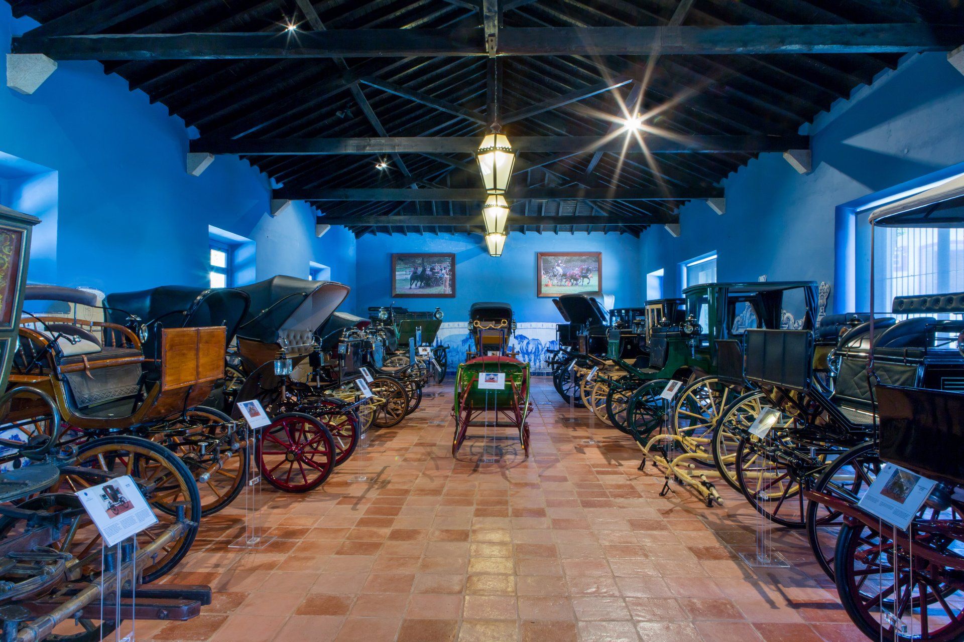 Museum exhibit of horse-drawn carriages on display. Blue walls, brown tiled floor, and dark wooden ceiling.