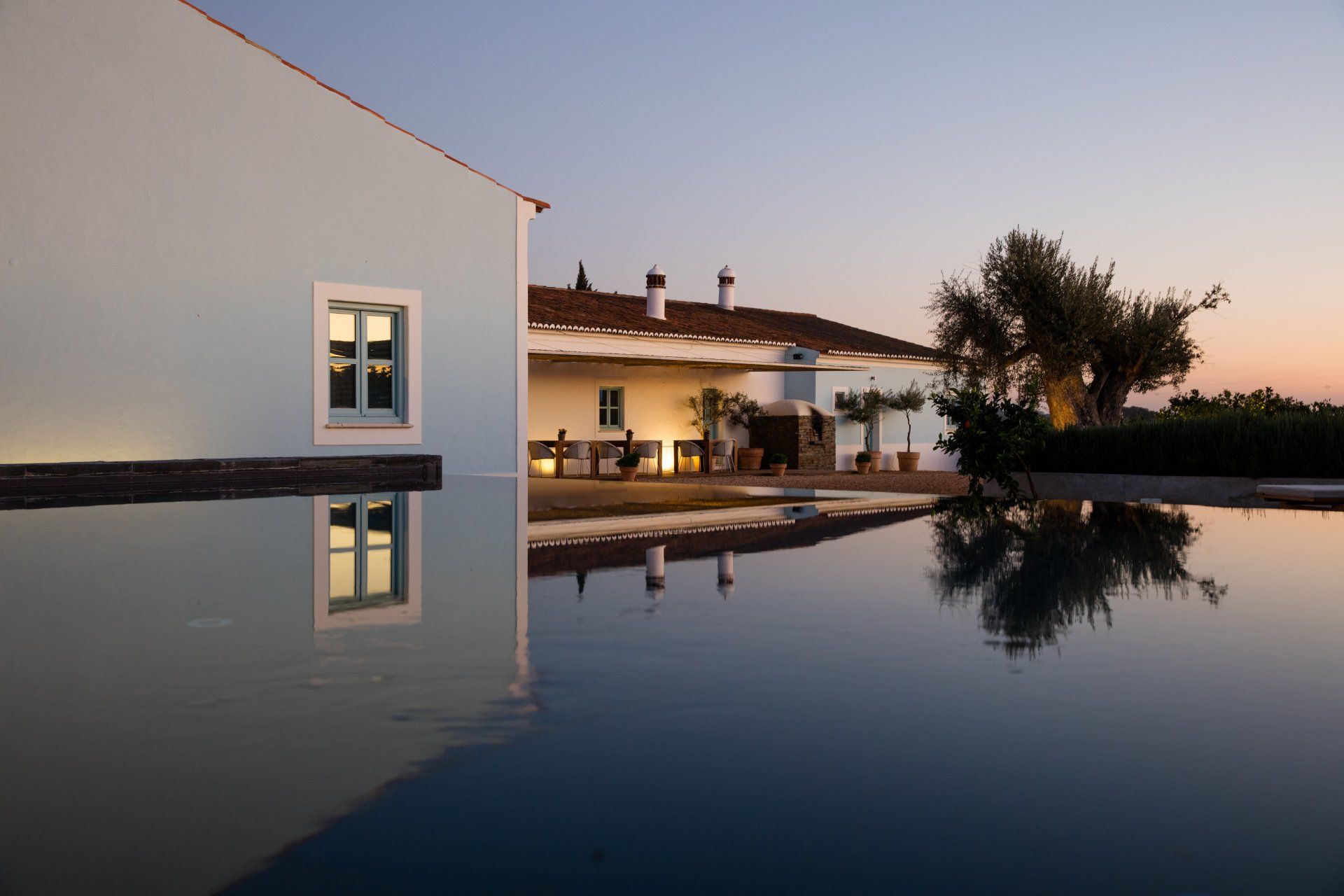 A house reflected in a pool at dusk, with pale blue walls and a white-roofed veranda.