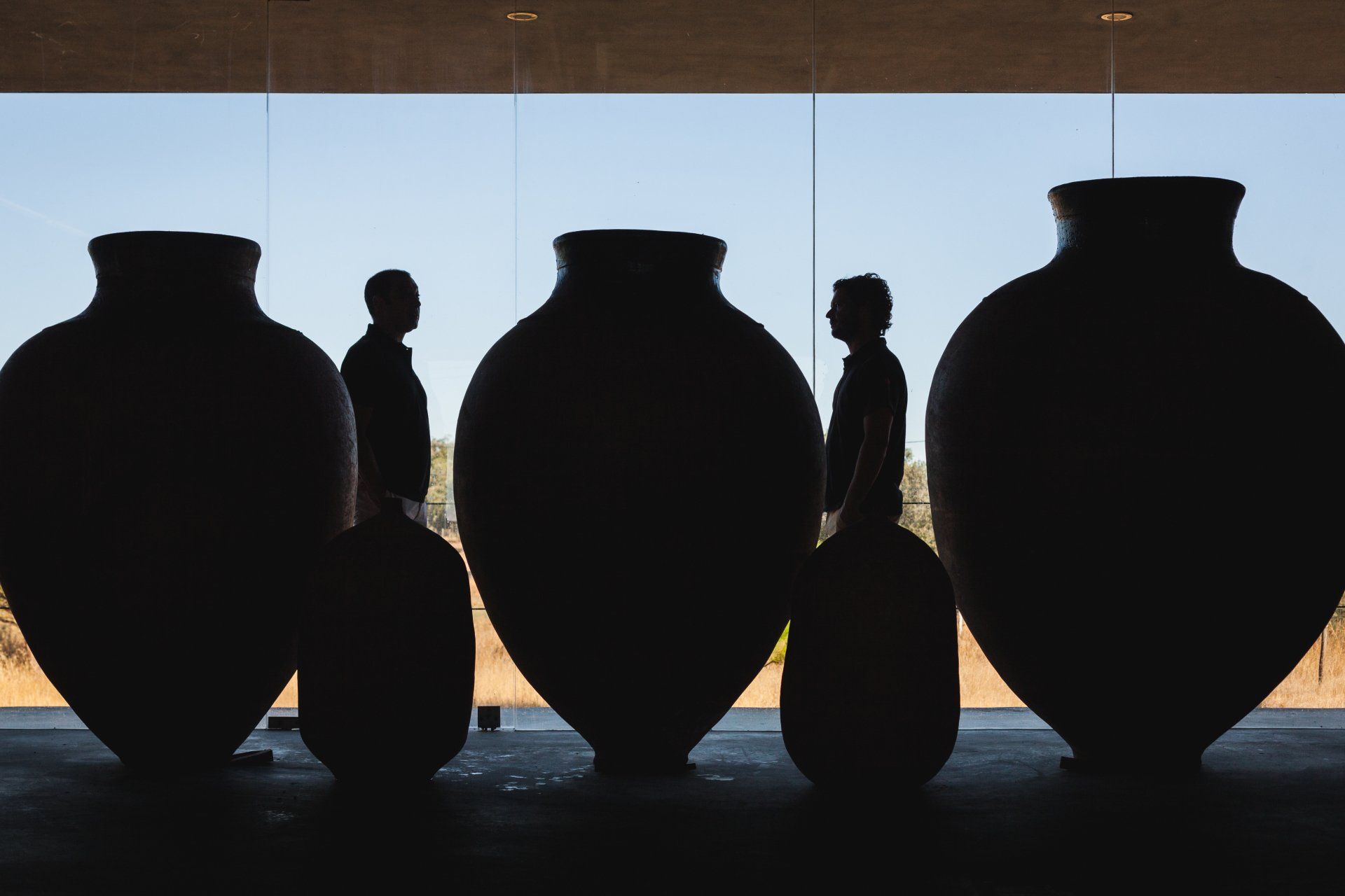 Silhouetted figures beside large, dark pottery against a glass wall; outdoor setting.