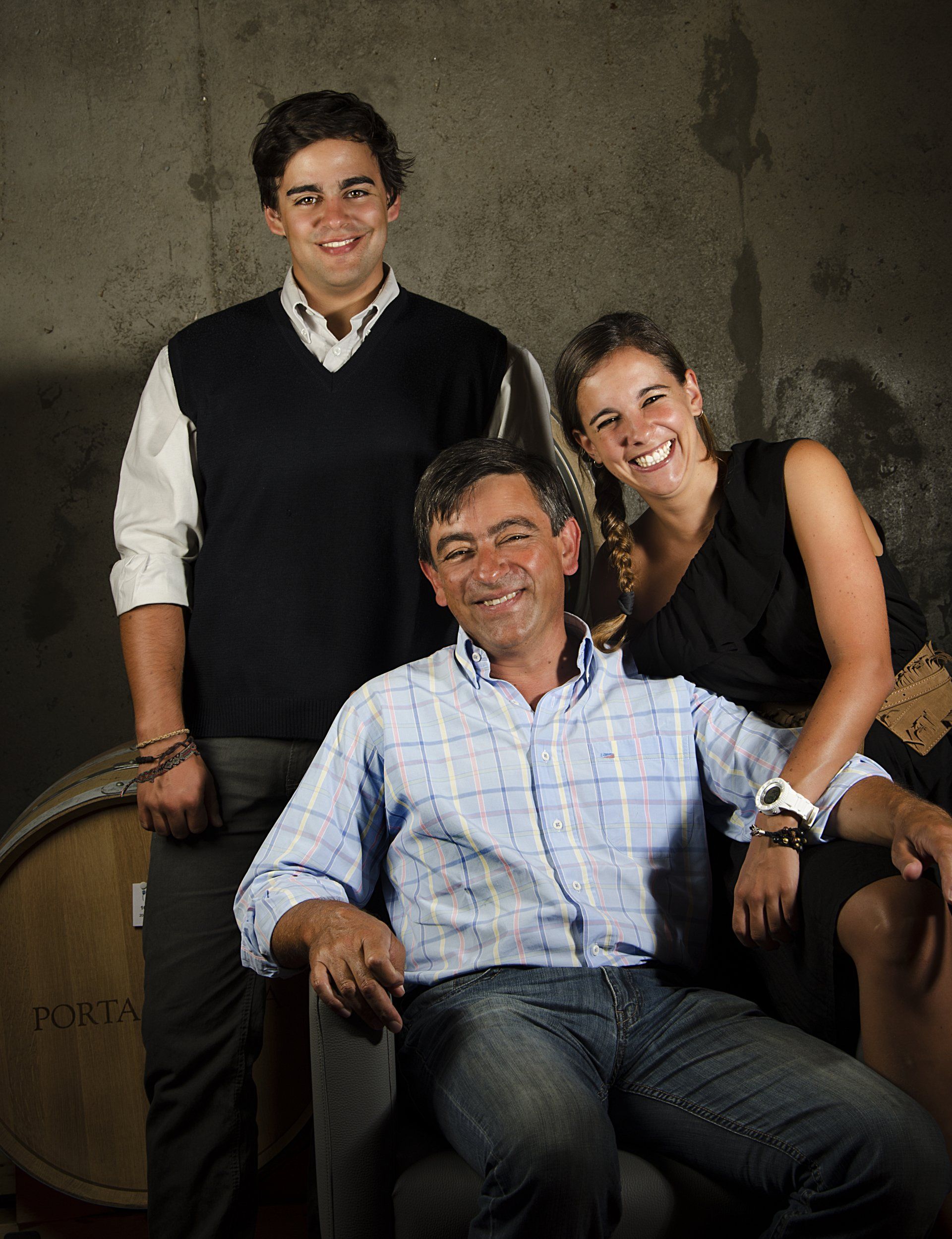 Family posing in front of a wine barrel; man in blue shirt seated, flanked by a young man and woman.