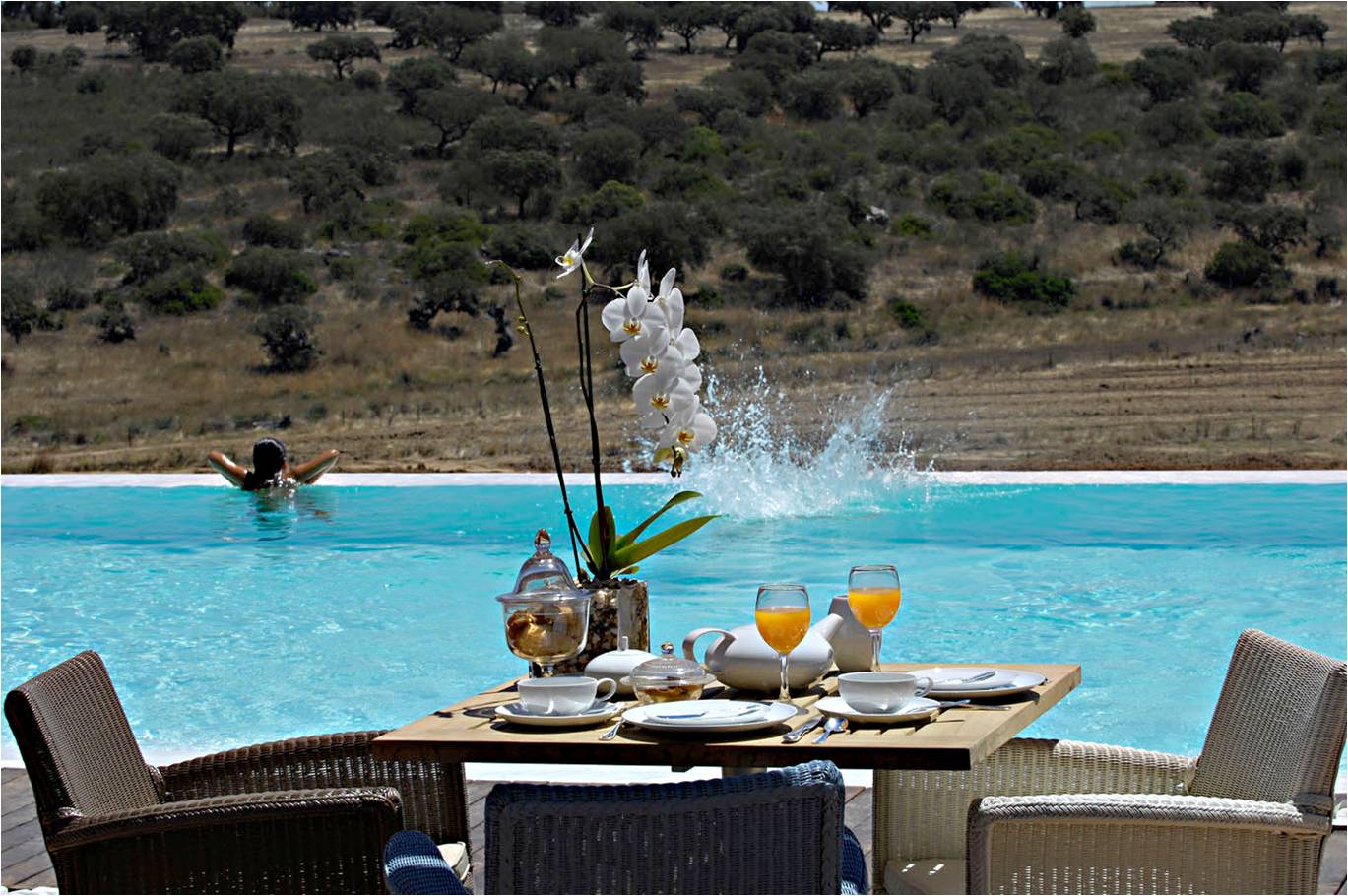 Table set for breakfast next to an infinity pool with someone swimming, olive trees in the background.