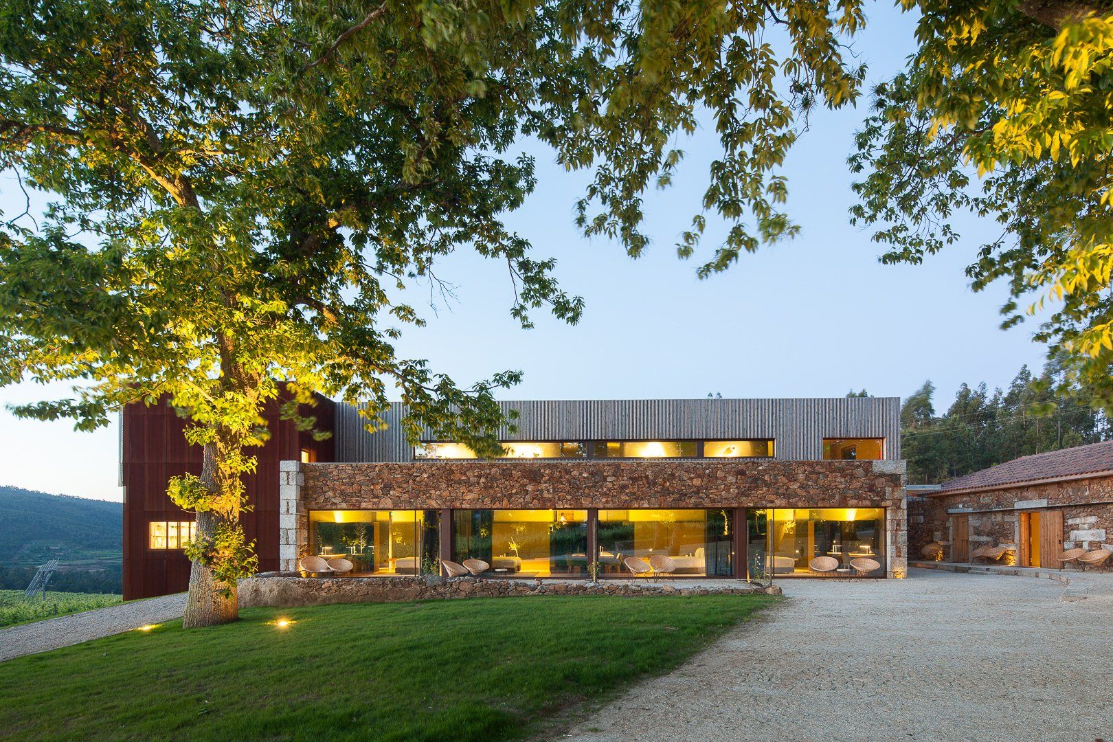 Modern building with glass walls, stone facade, and wooden upper section, framed by trees and green lawn.