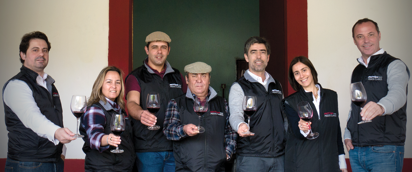 Group of people holding up glasses of red wine. They're standing in front of a doorway.