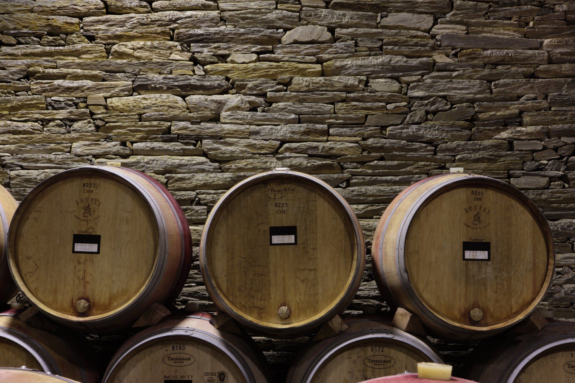 Wine barrels stacked in a cellar against a textured stone wall.