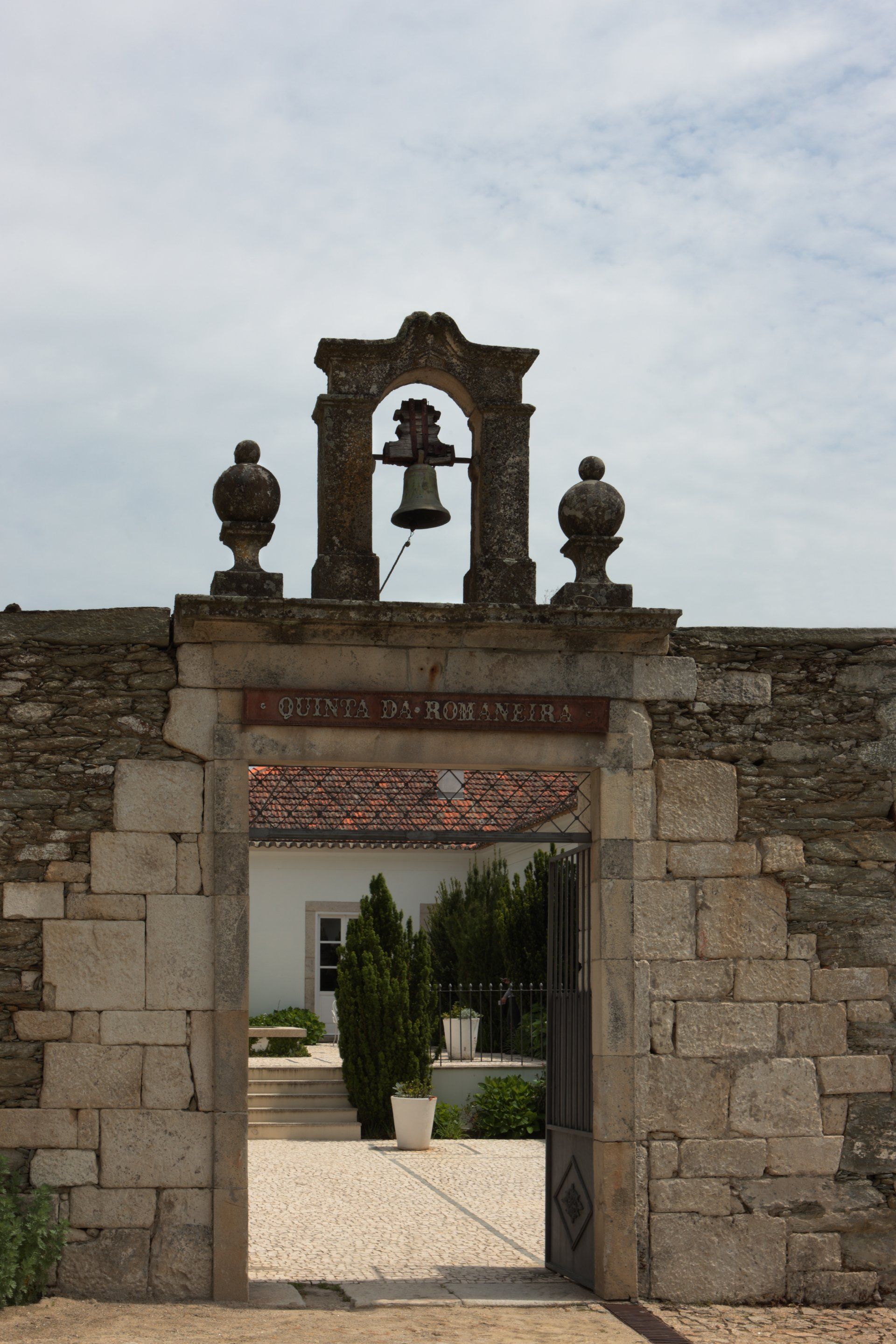 Stone archway with a bell, leading to a white building with red-tiled roof.